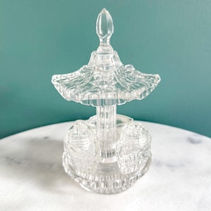 May include: Clear glass salt and pepper shaker set with a decorative, tiered design. The set includes two shakers and a central stand with a decorative top. The glass has a cut-glass appearance, adding to its vintage charm. The set is designed for tabletop use.