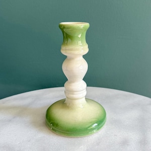 May include: A ceramic candlestick holder with a green and white ombre design. The base and top are green, while the middle section is white. The candlestick holder has a round base and a detailed, decorative design.