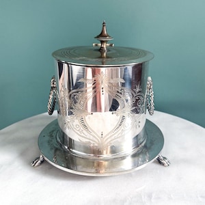 May include: A silver ice bucket with a lid and etched floral designs. The bucket has two chain handles and rests on a round base with claw feet. The lid is topped with a decorative finial. The bucket is approximately 15 cm in height.