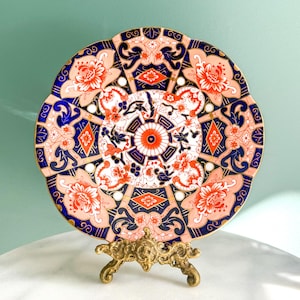 May include: A decorative plate with a floral design in shades of coral, navy blue, and white, displayed on a gold-coloured metal stand. The plate features intricate patterns and a scalloped edge, showcasing a vintage aesthetic.