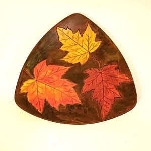 Peut inclure: Une assiette décorative triangulaire marron avec trois feuilles d'érable aux couleurs d'automne. Les feuilles sont peintes dans des tons de jaune, d'orange et de rouge, avec des motifs de veines détaillés. L'assiette a un aspect rustique et artisanal.
