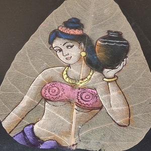 May include: A painting of a woman holding a pot, painted on a dried leaf. The woman is wearing a pink top, a purple skirt, and a gold necklace. The leaf is light brown and has a delicate vein pattern.