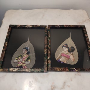 May include: Two framed art pieces featuring hand-painted images of women on dried leaves. The frames are black with a floral pattern and the leaves are a light brown colour. The women are wearing traditional Indian clothing and are depicted in different poses.