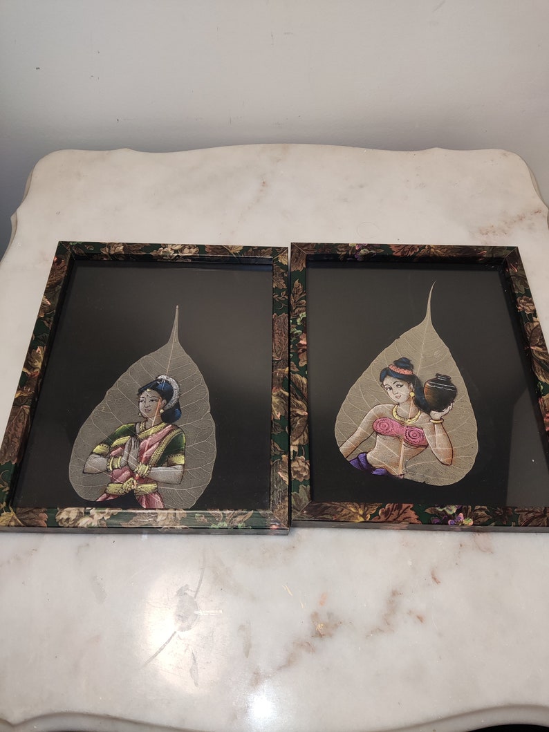 May include: Two framed art pieces featuring hand-painted images of women on dried leaves. The frames are black with a floral pattern and the leaves are a light brown colour. The women are wearing traditional Indian clothing and are depicted in different poses.