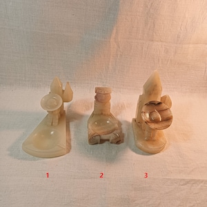 May include: Three carved stone figurines in shades of beige and brown. The figurines depict a person with a hat, a bottle, and a person with a round object. The objects are displayed on a white surface.