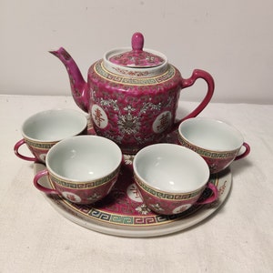 May include: A pink and white porcelain tea set with a teapot and four cups. The teapot has a floral design and the cups have a geometric design. The set is on a matching plate.