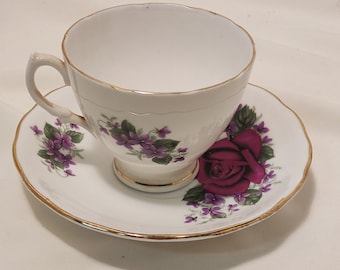 Può includere: Un set da tè bianco con bordo dorato. Il piattino ha una rosa rossa e fiori viola. La tazza da tè ha fiori viola.