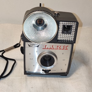 Könnte beinhalten: Vintage Imperial Lark Kamera mit schwarzem Gehäuse und silberner Frontplatte. Die Kamera hat einen Blitz, ein Objektiv und den roten Schriftzug "LARK". Mit schwarzem Riemen. Hergestellt in den USA.