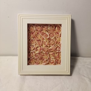 May include: A square, off-white framed artwork filled with numerous small, light pink and cream-colored roses. The frame has a simple, classic design. The roses are densely packed, creating a textured, floral display.