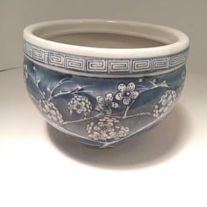 Pode incluir: Um vaso redondo de cerâmica azul e branco com um design floral. O vaso apresenta uma borda branca com um padrão geométrico e um corpo azul adornado com ramos de flores de cerejeira brancas. Ideal para pequenas plantas ou suculentas.