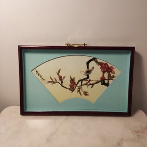 May include: A framed wall art piece with a light blue background and a yellow fan-shaped design. The fan features a bird and a branch with red flowers made of shell pieces.