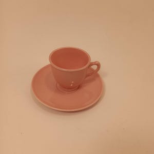 Puede incluir: Un juego de taza y platillo de cerámica rosa. La taza tiene un asa y se apoya en un platillo a juego. El juego es de color rosa suave y liso, y probablemente sea vintage o retro.
