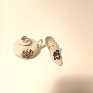 May include: Two small, cream-colored ceramic decorative items. One is a round oil lamp with a handle and a gold rim. The other is a miniature shoe. Both items feature a colorful coat of arms design.