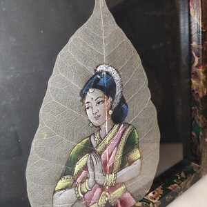May include: A hand-painted depiction of a woman in traditional Indian clothing, with her hands clasped in a prayer position, is painted on a delicate, translucent leaf. The leaf is framed in a dark wood frame with a floral design.