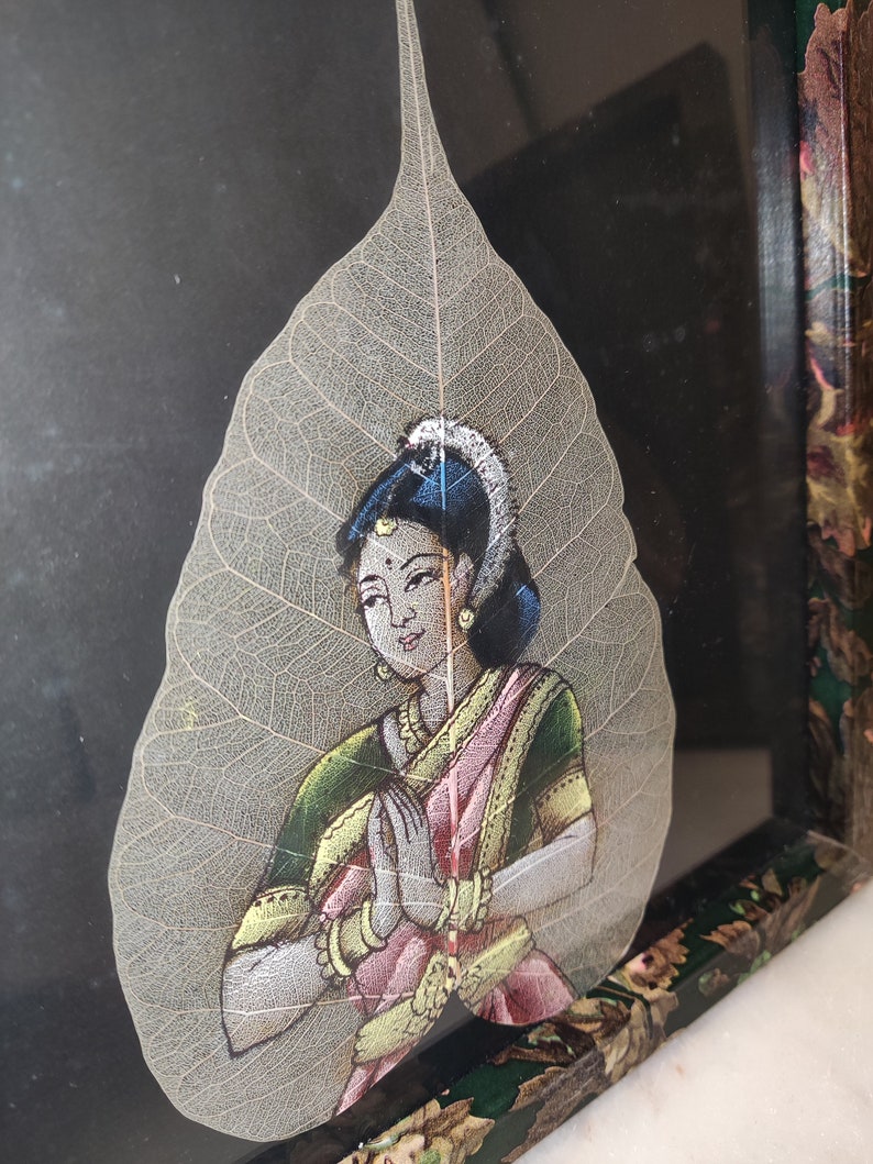 May include: A hand-painted depiction of a woman in traditional Indian clothing, with her hands clasped in a prayer position, is painted on a delicate, translucent leaf. The leaf is framed in a dark wood frame with a floral design.