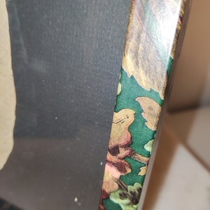 May include: A close-up of the edge of a picture frame with a green background and a floral pattern. The frame is made of wood and has a gold-coloured finish.