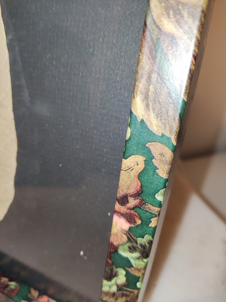 May include: A close-up of the edge of a picture frame with a green background and a floral pattern. The frame is made of wood and has a gold-coloured finish.