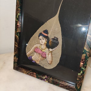 May include: A framed artwork featuring a woman painted on a dried leaf. The woman is holding a pot and wearing a pink top and a gold necklace. The background is black.