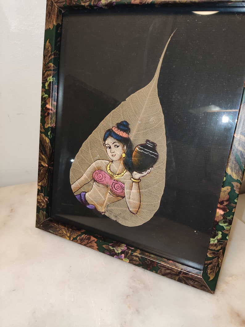 May include: A framed artwork featuring a woman painted on a dried leaf. The woman is holding a pot and wearing a pink top and a gold necklace. The background is black.