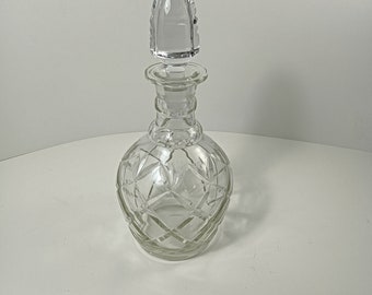 May include: A clear glass decanter with a diamond-cut pattern and a matching stopper. The decanter has a round body and a long neck, and the stopper is shaped like a teardrop. The glass is transparent, allowing the contents to be visible.