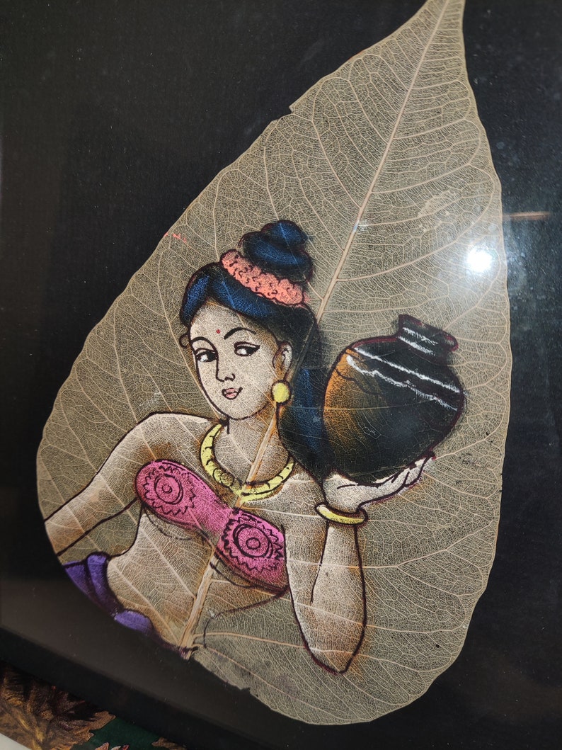 May include: A painting of a woman carrying a pot on a dried leaf. The woman is wearing a pink top, a gold necklace, and a blue head scarf. The leaf is beige and has a detailed vein pattern.
