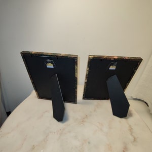 May include: Two black picture frames with a floral patterned border. The frames are standing upright on a white surface.