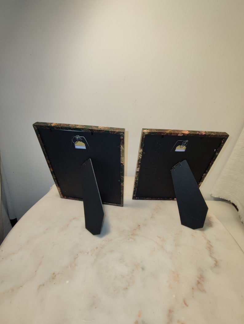 May include: Two black picture frames with a floral patterned border. The frames are standing upright on a white surface.
