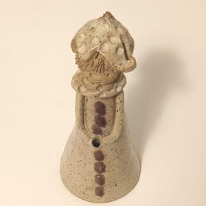 May include: A handcrafted ceramic figurine, featuring a mushroom-style hat and a long dress. The beige figure has brown detailing, including spots on the hat and a series of markings down the front. The design is whimsical and artistic, with a unique, handcrafted aesthetic.