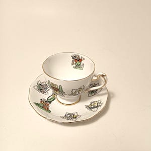 May include: A white teacup and saucer set with gold trim and colourful illustrations. The teacup has a handle and a pedestal base. The saucer features multiple illustrations. The teacup has a cartoon character illustration.