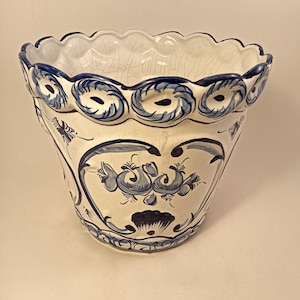 May include: A white ceramic planter with blue floral designs and a scalloped rim. The planter features hand-painted blue swirls and floral motifs. The interior is white with visible crackling. A decorative piece for indoor or outdoor use.