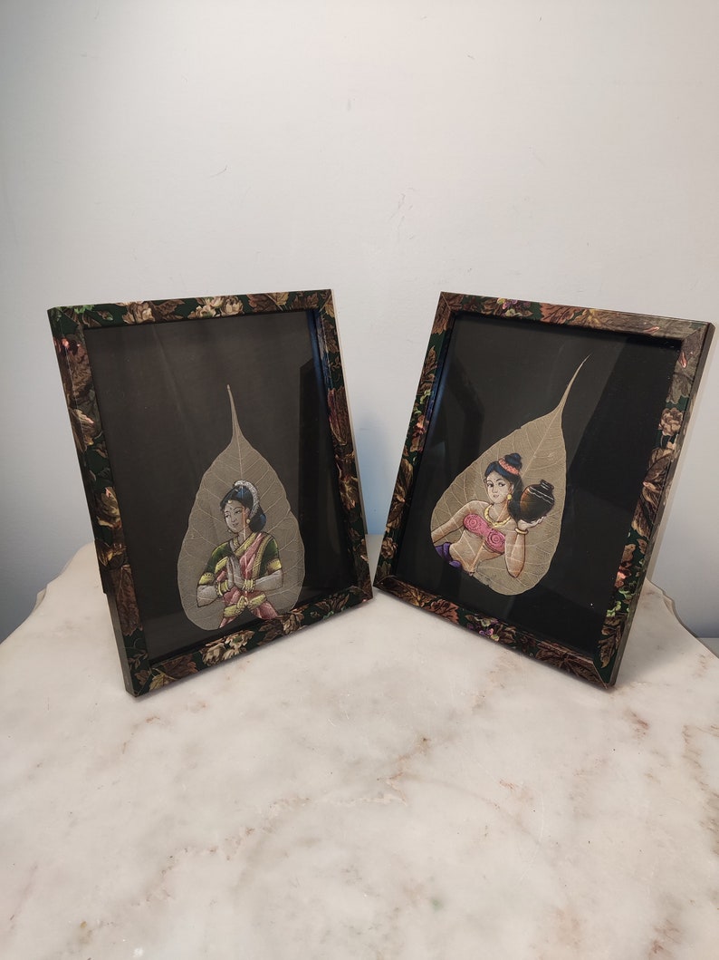 May include: Two framed paintings of women in traditional Indian clothing, painted on dried leaves. The frames are decorated with a floral pattern in shades of green and brown.