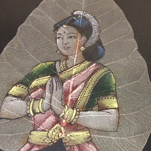 May include: A woman in a pink and green sari with gold jewellery is painted on a translucent leaf. The woman is holding her hands together in a prayer position.