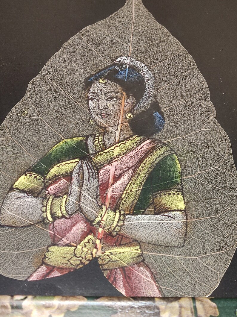 May include: A woman in a pink and green sari with gold jewellery is painted on a translucent leaf. The woman is holding her hands together in a prayer position.