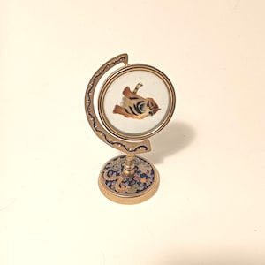 May include: A small, decorative brass globe-shaped stand with a circular frame holding a detailed embroidered image of a tiger. The base features a blue background with colorful floral and dragon patterns.