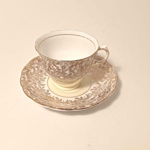 May include: A cream-coloured teacup and saucer set with a gold floral design and gold edging. The teacup has a handle and sits on a matching saucer. The set is likely made of porcelain, a classic for afternoon tea.