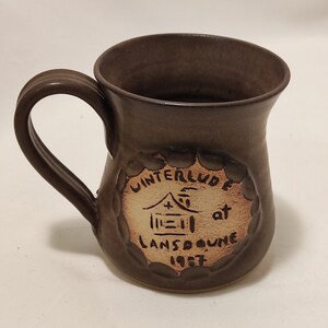 May include: A brown ceramic mug with a handle. The mug has a white design on the front that says "Winterlude at Lansdowne 1987".