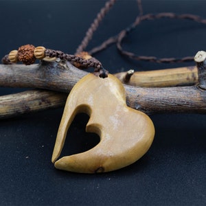 Puede incluir: Un colgante de madera en forma de corazón con un diseño de corazón recortado, colgando de un cordón marrón con cuentas marrones. El colgante está sobre una rama marrón.