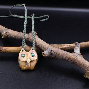 Könnte beinhalten: Ein brauner Eulenaanhänger mit türkisfarbenen Augen hängt an einer grünen Schnurkette. Die Kette liegt über zwei braunen Ästen.