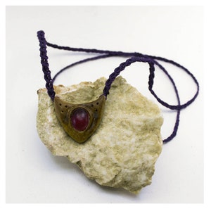 Puede incluir: Un colgante de oro con una piedra preciosa roja engastada en un marco triangular que cuelga de un cordón morado. El colgante descansa sobre una pieza de roca rugosa de color claro.