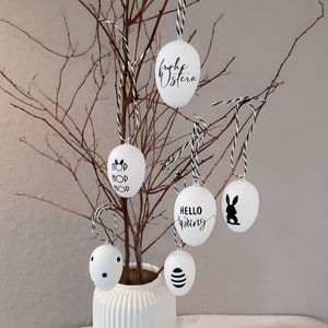 Könnte beinhalten: Fünf weiße Ostereier hängen an einem kahlen Zweig in einer weißen Vase. Die Eier sind mit schwarzer Schrift und Designs verziert, darunter "Frohe Ostern", "Hop Hop Hop", "Hallo Frühling" und ein Kaninchen.