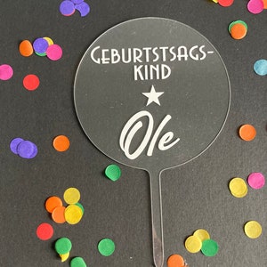 Könnte beinhalten: Klarer Acryl-Kuchentopper mit dem Text "Geburtstagskind" und einem Stern über dem Wort "Ole". Der Topper ist von buntem Konfetti umgeben.