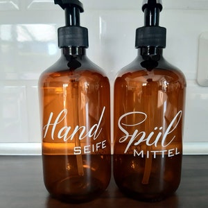 Könnte beinhalten: Zwei bernsteinfarbene Glasflaschen mit schwarzen Pumpen. Die Flaschen sind mit "Hand Seife" und "Spülmittel" in weißer Schrift beschriftet.