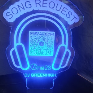 Puede incluir: Un letrero de acrílico azul y blanco con el texto "SONG REQUEST" en una fuente curvada. El letrero presenta una imagen estilizada de auriculares con un código QR en el interior. El texto "One28" y "DJ GREENHIGH" también están incluidos en el letrero.
