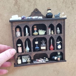 Pode incluir: Uma prateleira de madeira castanha escura com compartimentos arqueados, cheia de garrafas de boticário em miniatura, frascos e objetos decorativos. A prateleira tem um topo decorativo e está colocada sobre um fundo neutro. As garrafas são de várias formas e cores.