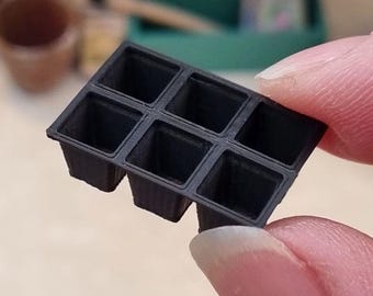 Pode incluir: Uma pequena bandeja de plástico preto para sementes com seis células quadradas. A bandeja é segurada entre dois dedos, com outros materiais de jardinagem visíveis no fundo desfocado.