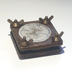 May include: A wooden square box with a decorative tile in the center. The box has four brass wing nuts on the corners.