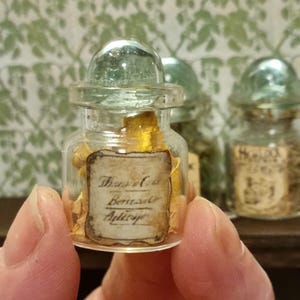 May include: A small glass bottle with a glass stopper filled with yellow and gold flakes. The bottle has a label that reads "Thymol et Bergamote Apéritif".