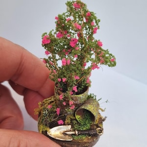 以下が含まれることがあります： ピンクの花と緑の葉を持つ鉢植えの植物を特徴とするミニチュアガーデンシーン。 小さなシャベルと、魅力的な手作りの美学を作り出す、風化した苔で覆われたベースが含まれています。 全体的なテーマは、ガーデニングとミニチュアです。