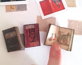 May include: Three miniature books, two closed and one open. The closed books have dark brown and red covers with text. The open book displays vintage-style illustrations of figures in a garden setting on its pages, showing detailed printed imagery.
