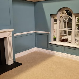 May include: A dollhouse room with blue walls, white trim, and a white fireplace. The room has a large arched window with white trim and a view of a garden.